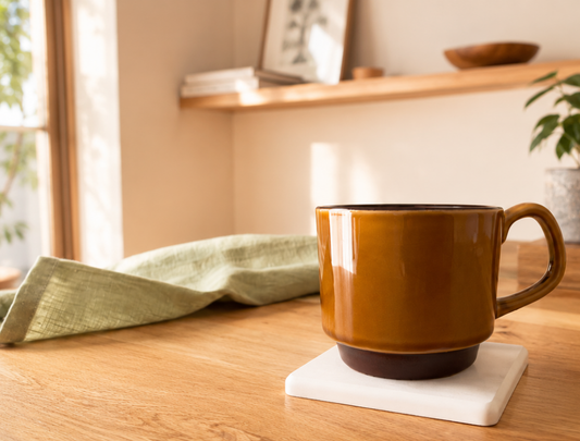 Brown ceramic mug on a white coaster on a wooden surface with a window and shelves in the background.